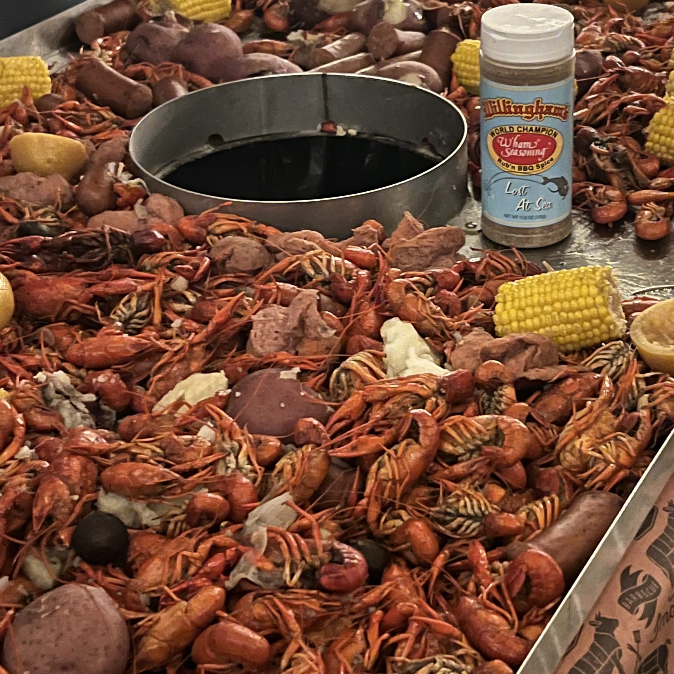 Pile of boiled crawfish with corn on the cob and potatoes, plus a spice bottle on a seafood boil tray.